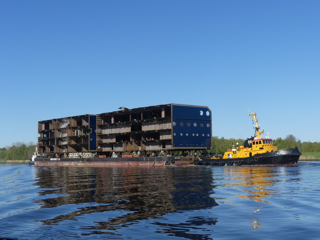 Unfinished multi-deck ship sections on a transport barge beside a yellow tug in the Kiel Canal.