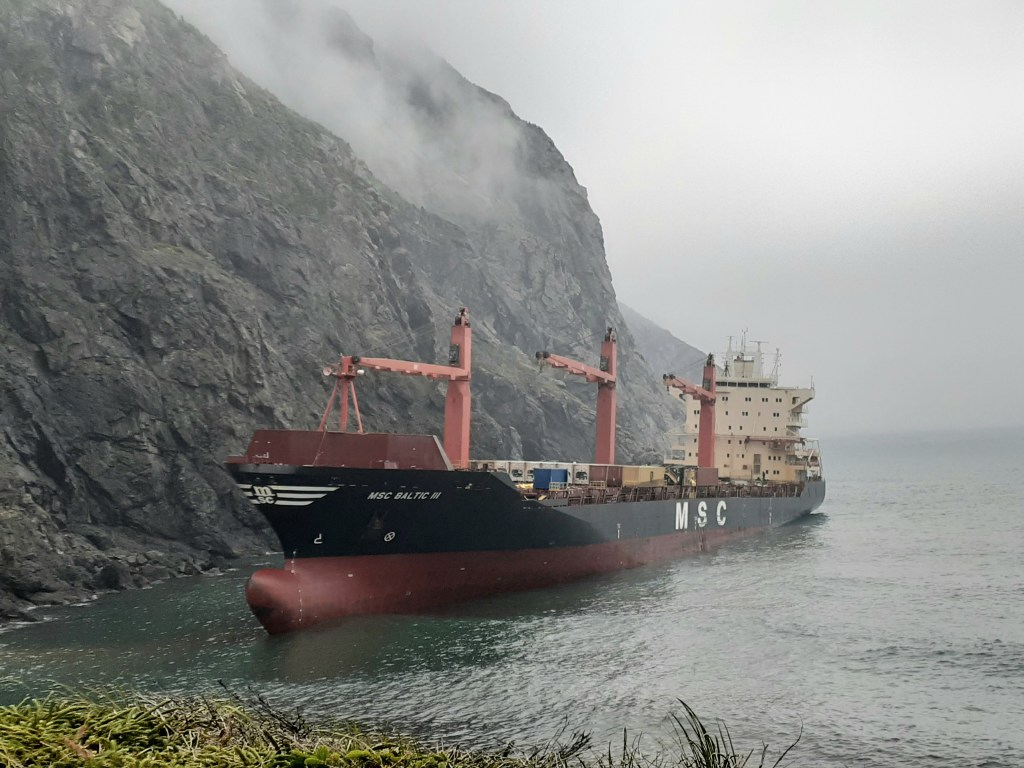 MSC Baltic III aground beside steep cliffs, with its bow name, three deck cranes and aft superstructure visible.